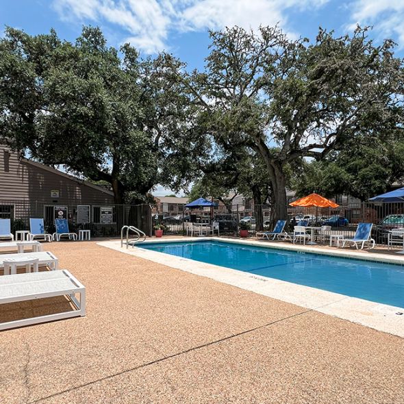 The pool at Autumn Chase Apartments in San Marcos, Texas, offers a relaxing setting with lounge chairs, umbrellas, and shaded areas surrounded by mature trees